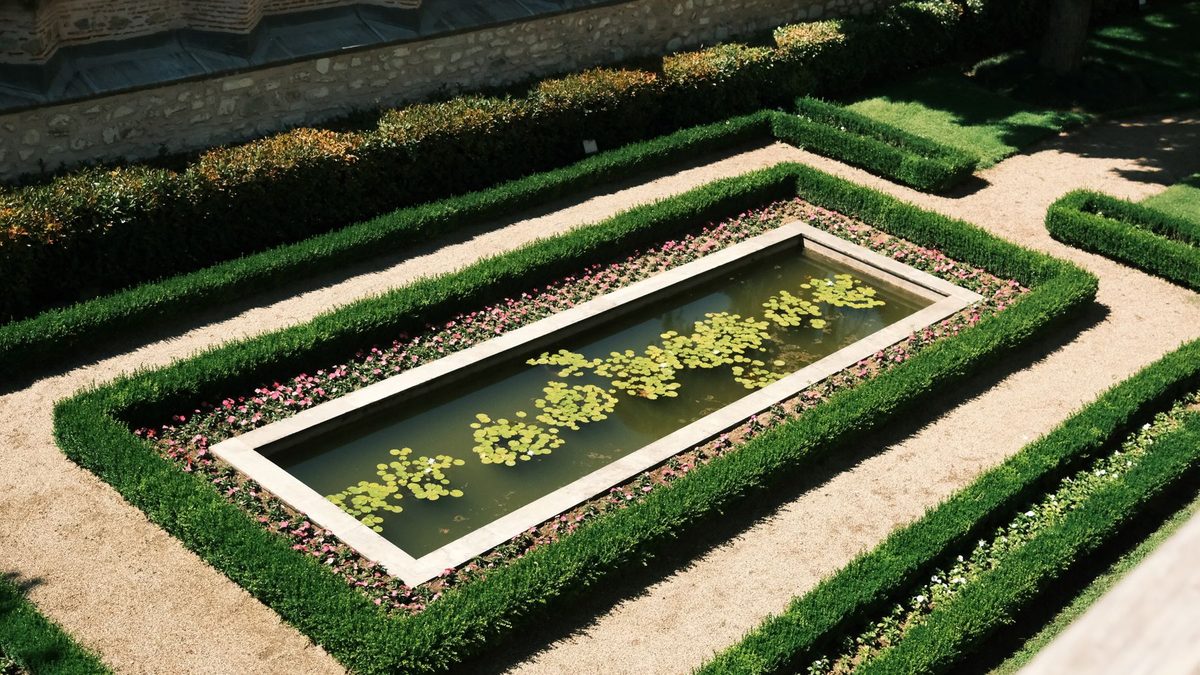 Elevated view of a rectangular lily pond framed by concentric clipped boxwood hedges, bands of pink and white bedding flowers and pale gravel paths