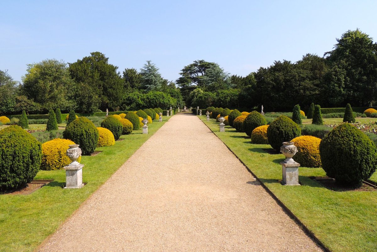 A wide gravel allée flanked by alternating dark green and golden clipped shrubs, carved stone urns on plinths and strips of mown lawn